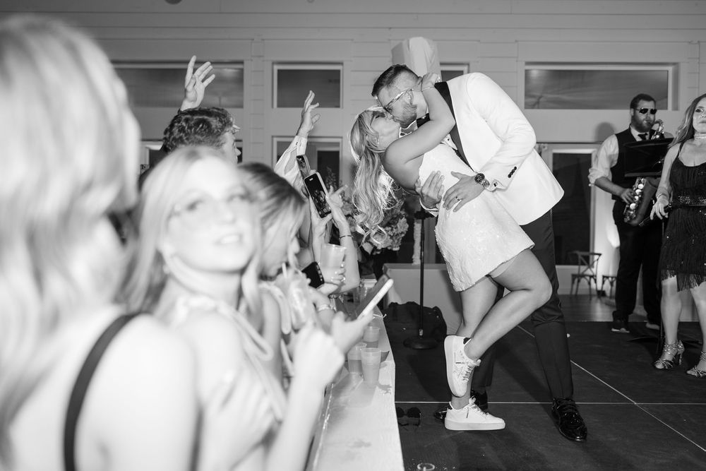 Black and white photo of bride and groom kissing on dance floor surrounded by cheering wedding guests.