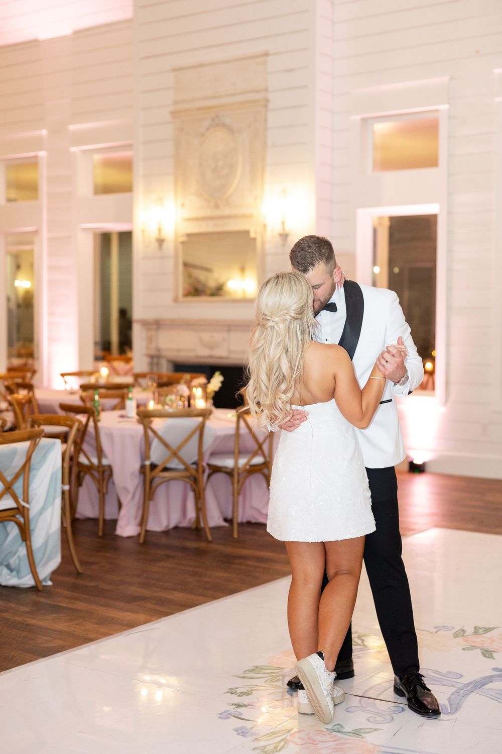 Bride in short white dress and groom in white tuxedo sharing first dance in elegant candlelit reception.