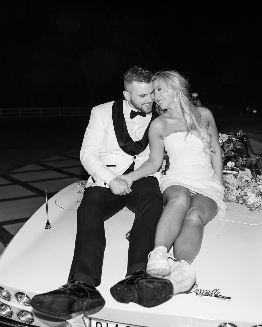 Black and white photo of bride and groom sitting on vintage car hood at night wedding.