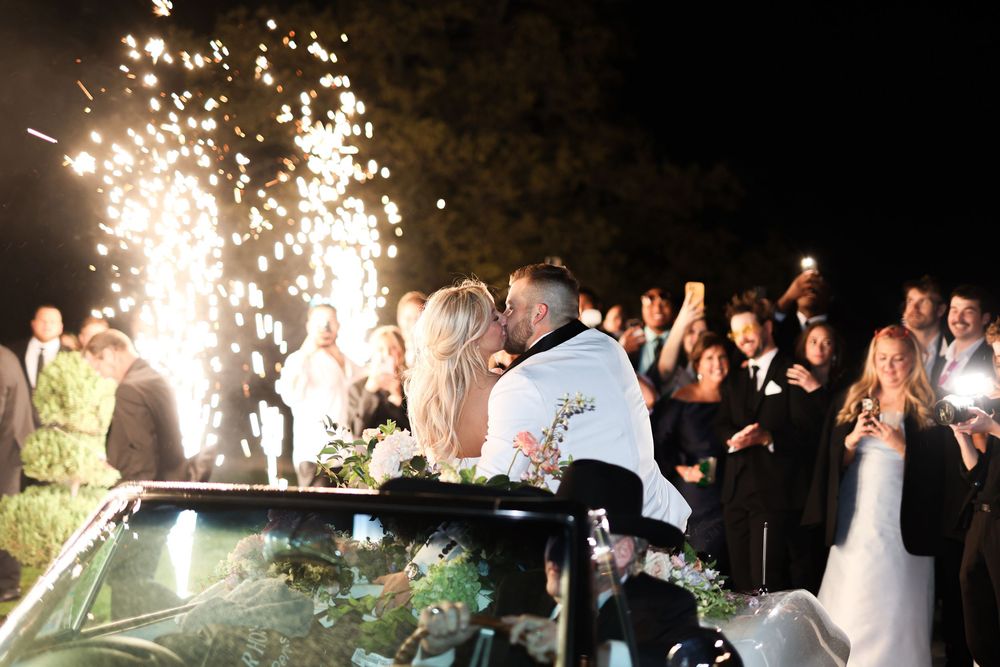 Couple shares kiss beside classic car with sparkler send-off at outdoor wedding reception.