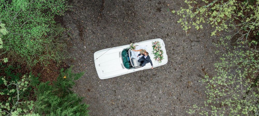 Aerial drone view of bride and groom in white convertible car surrounded by greenery.