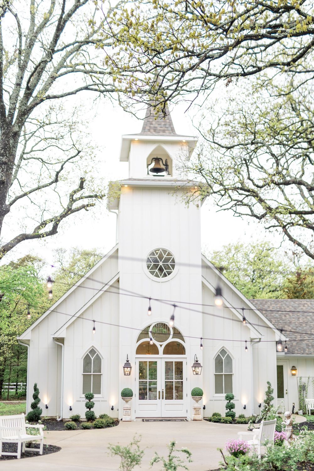 Charming white wedding chapel with bell tower, string lights, arched windows, and lush spring trees in romantic outdoor setting.