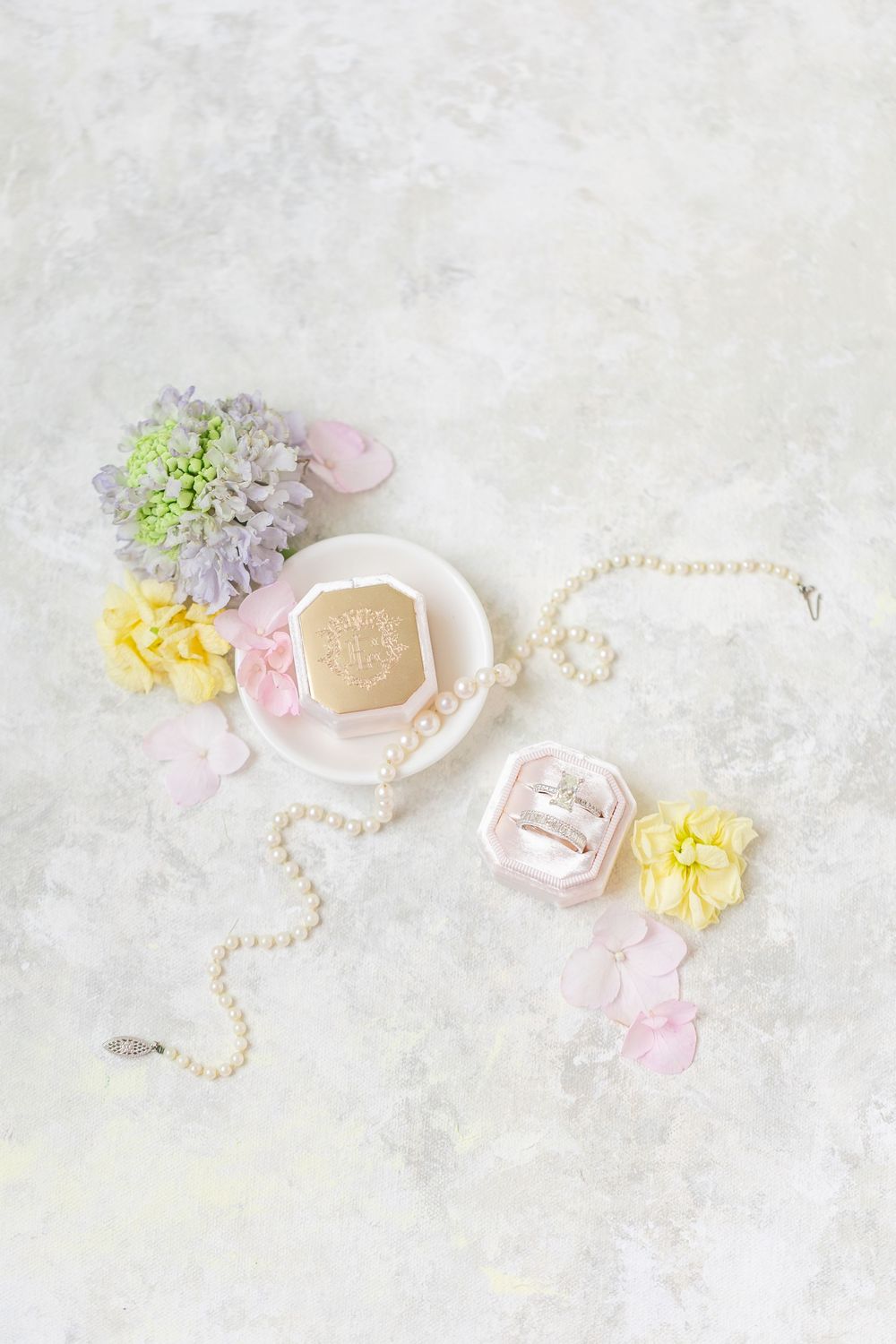 Romantic bridal flat lay with pearl necklace, diamond rings in pink velvet box, gold monogram box, and pastel spring flowers.