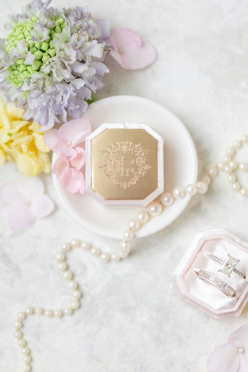 Elegant bridal detail flat lay featuring gold monogram ring box, pearl necklace, diamond rings, and soft pastel florals.