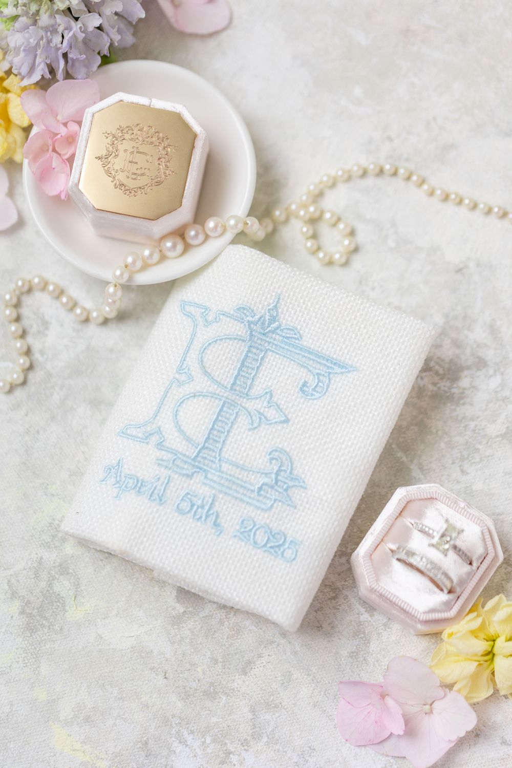 Bridal flat lay with embroidered monogram vow book dated April 5th 2025, pearl necklace, diamond ring, and pastel flowers.
