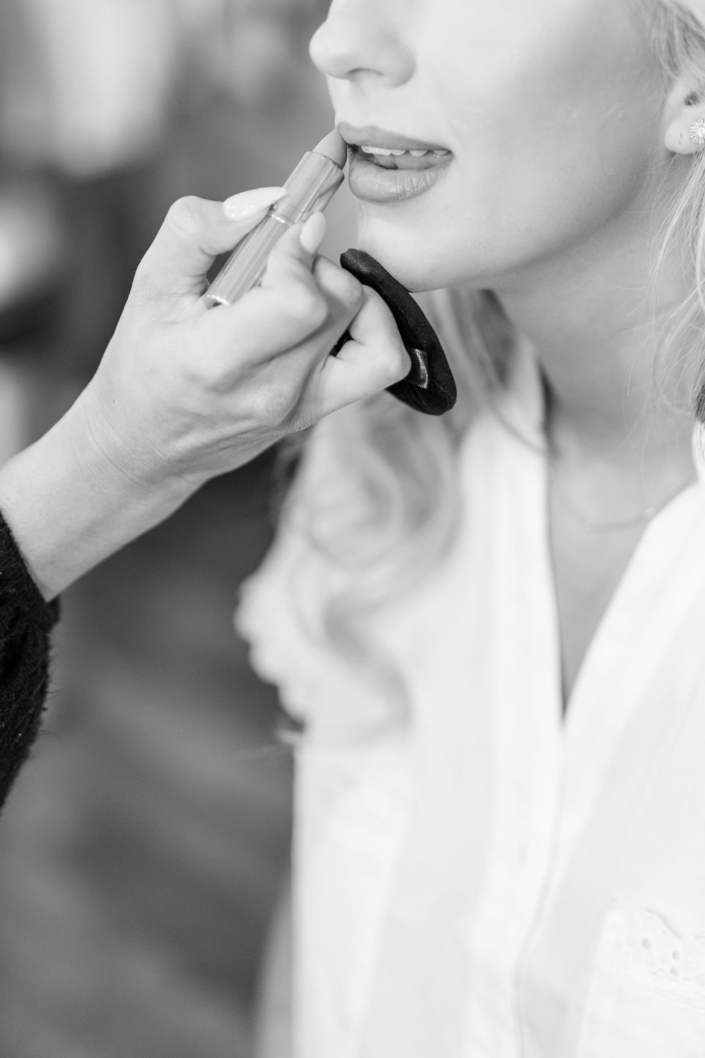 Black and white close-up of makeup artist applying lipstick to blonde bride in white robe during wedding morning prep.