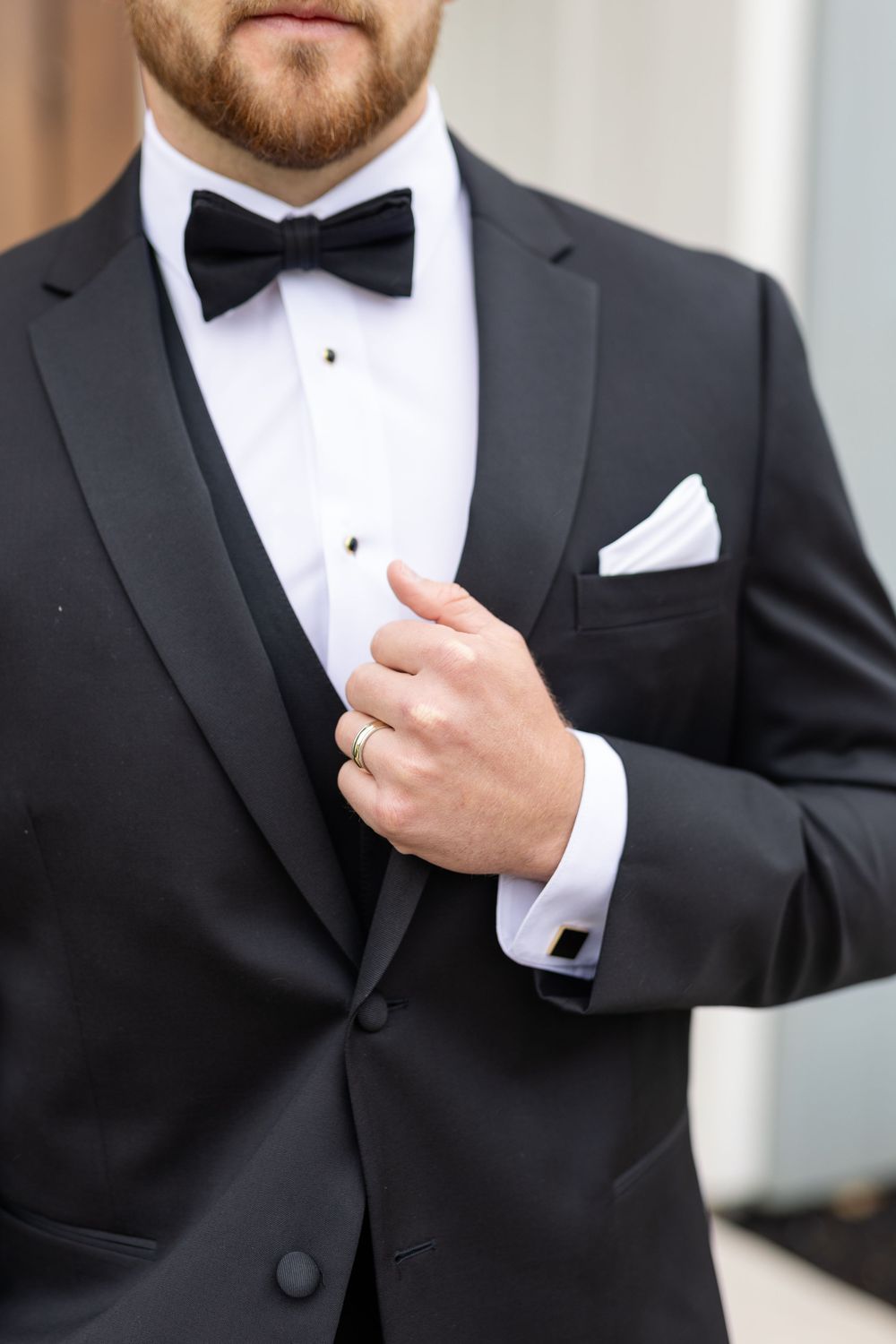 Groom in black tuxedo with bow tie and white pocket square adjusting his jacket lapel.