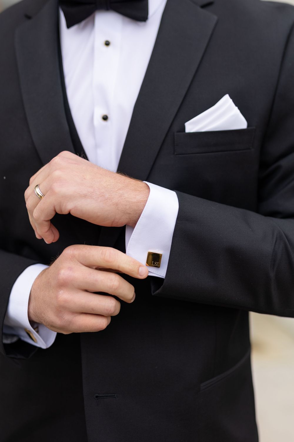 Groom in black tuxedo fastening gold cufflinks on white dress shirt sleeve.