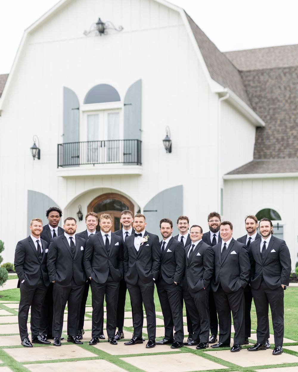 Large groomsmen party in matching black tuxedos posing in front of white barn wedding venue.