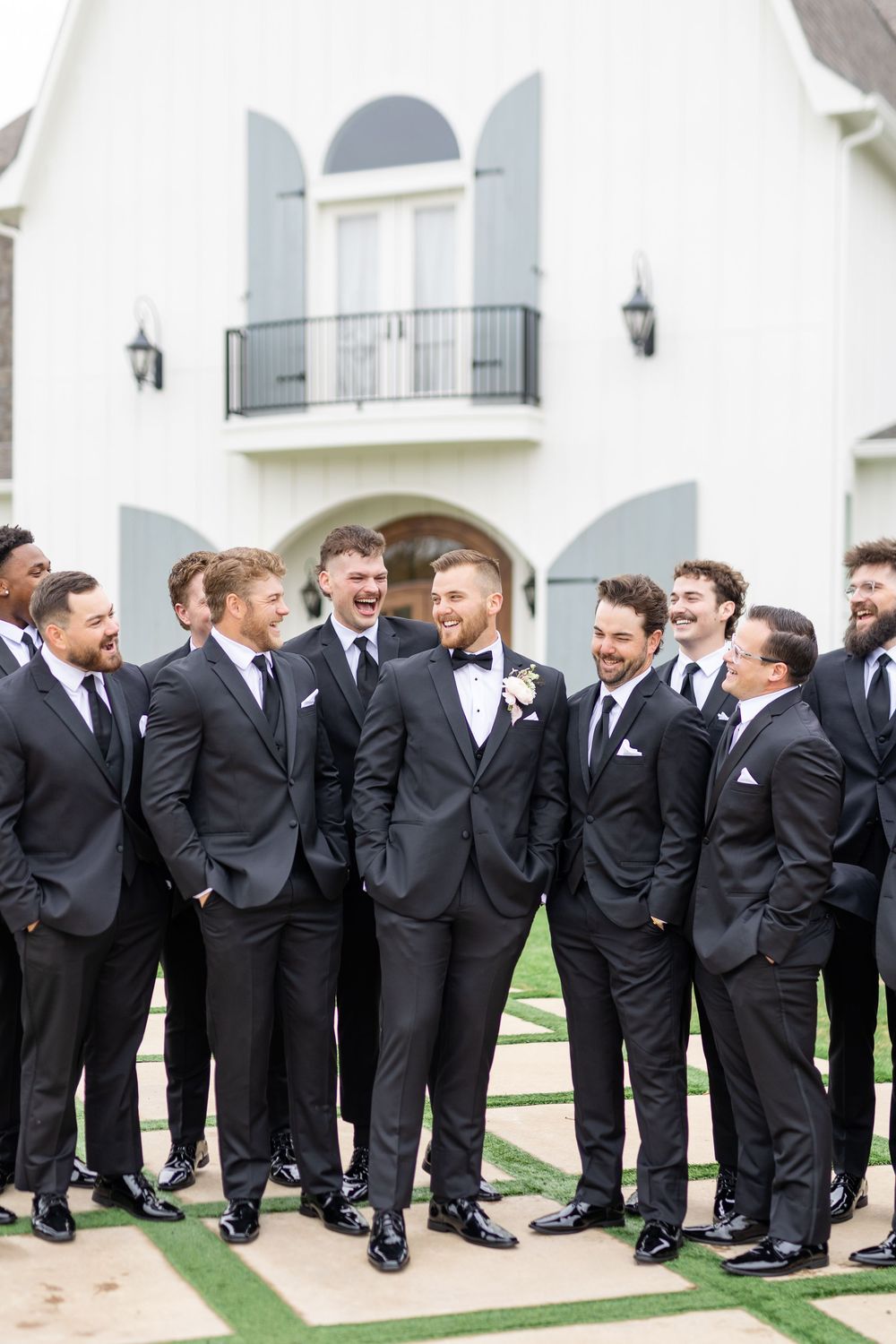Groom with boutonniere laughing with groomsmen in black tuxedos outside elegant white barn venue.