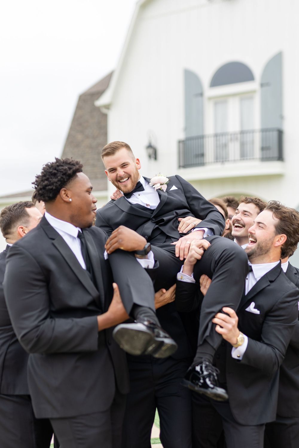 Groomsmen joyfully lifting smiling groom in black tuxedo outside white barn wedding venue.