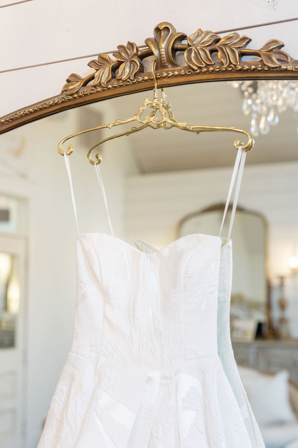 Strapless white lace wedding dress hanging on decorative gold hanger over ornate vintage mirror.