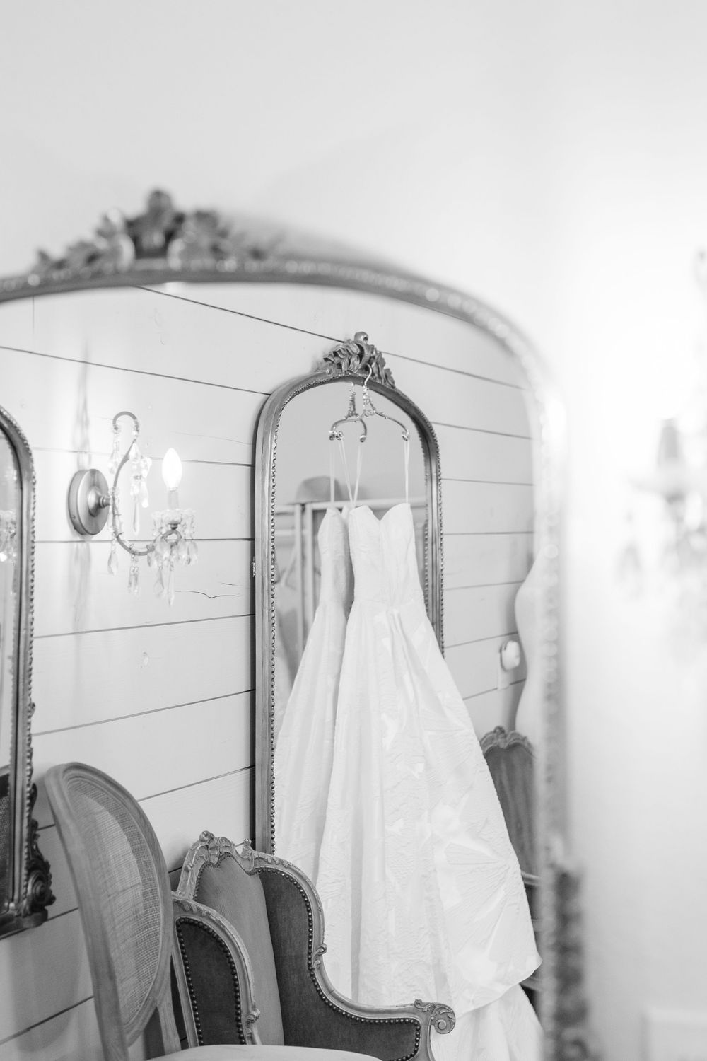 Black and white photo of strapless white wedding gown hanging on ornate gold mirror.