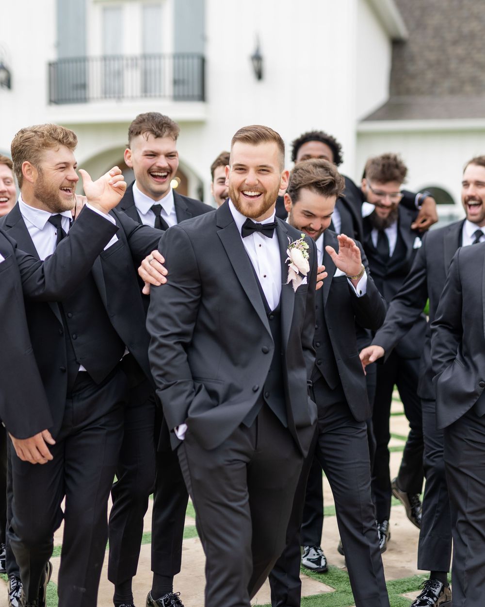 Smiling groom in black tuxedo walking with laughing groomsmen outside white barn wedding venue.