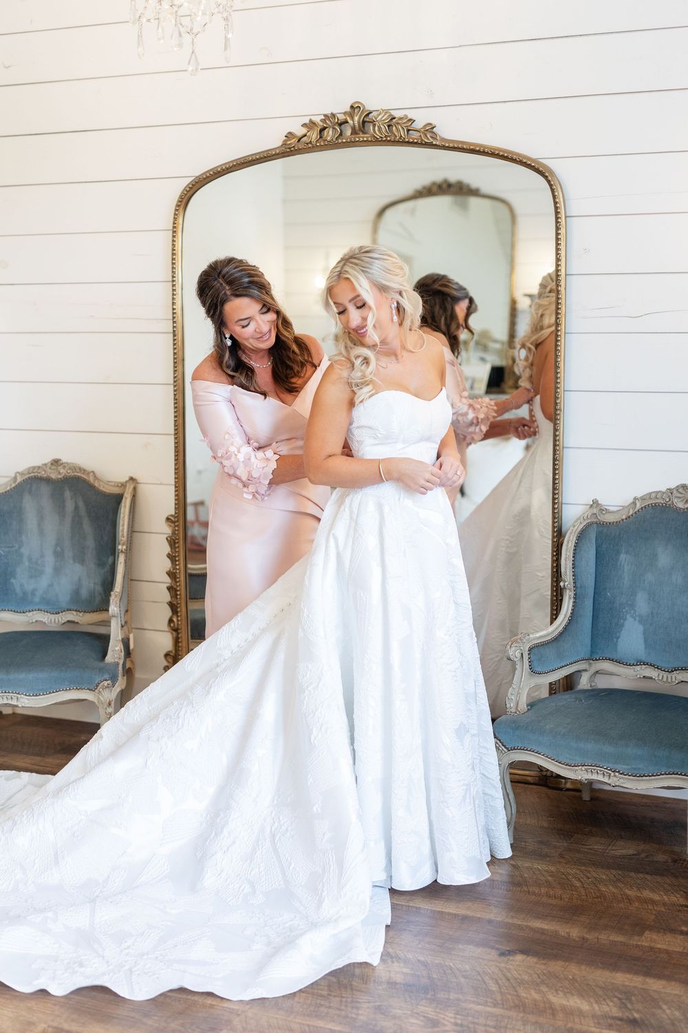 Bride in strapless white ball gown being helped by bridesmaid in front of large gold mirror.