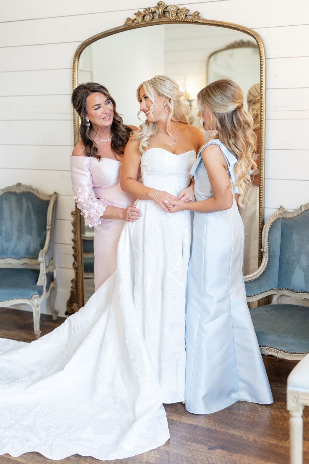 Bride in white gown with two bridesmaids in pink and blue dresses preparing in front of ornate gold mirror.