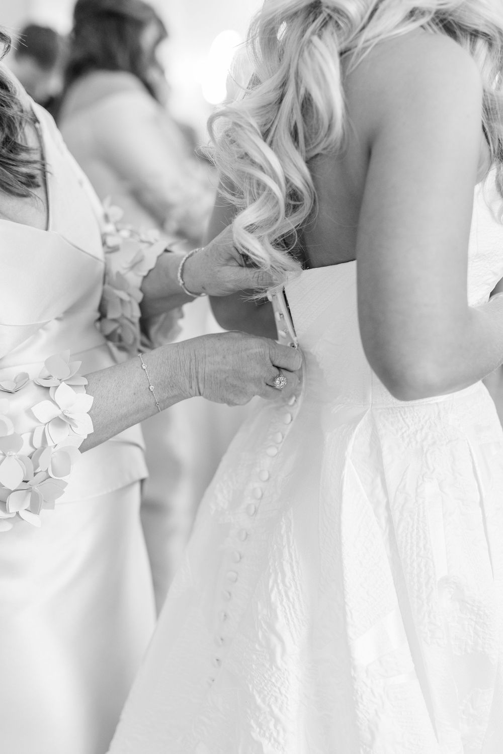 Black and white photo of bridesmaids buttoning the back of a brides elegant white wedding gown.
