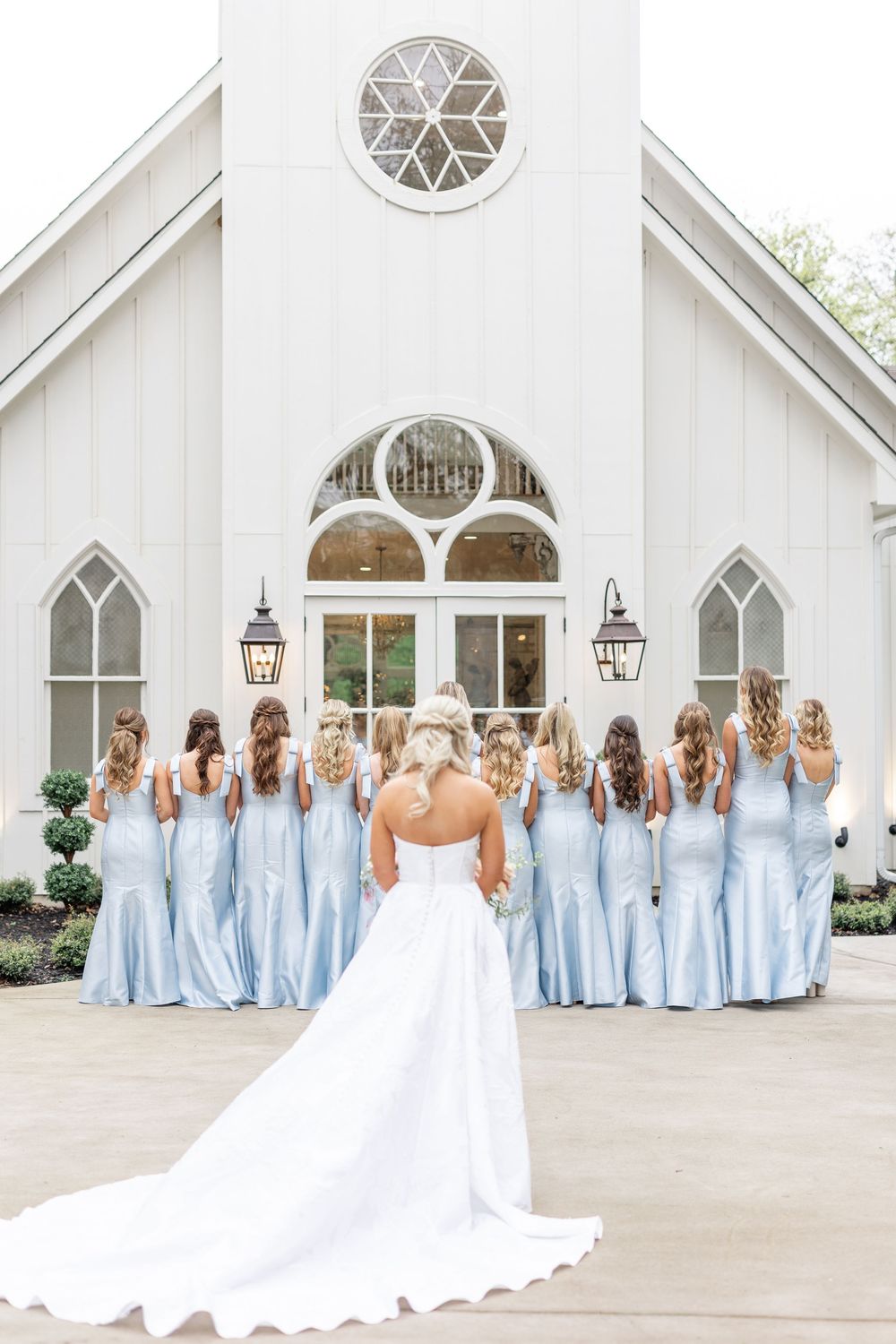 Bride in white gown with train faces bridesmaids in light blue dresses outside white chapel.