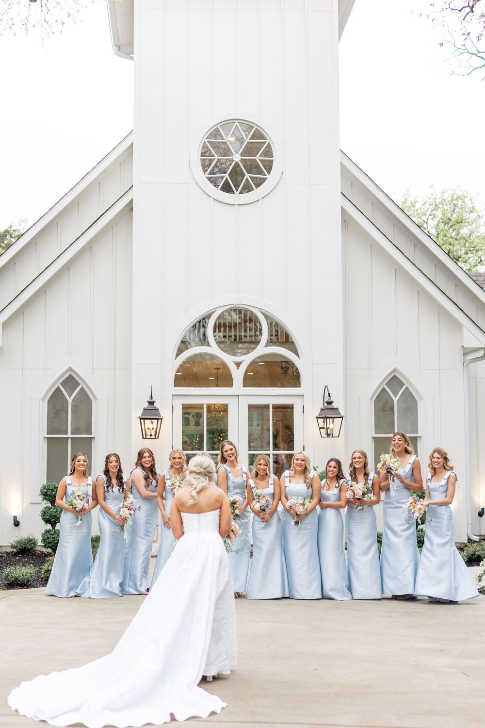Bride in white gown with train faces smiling bridesmaids in light blue dresses holding bouquets outside chapel.