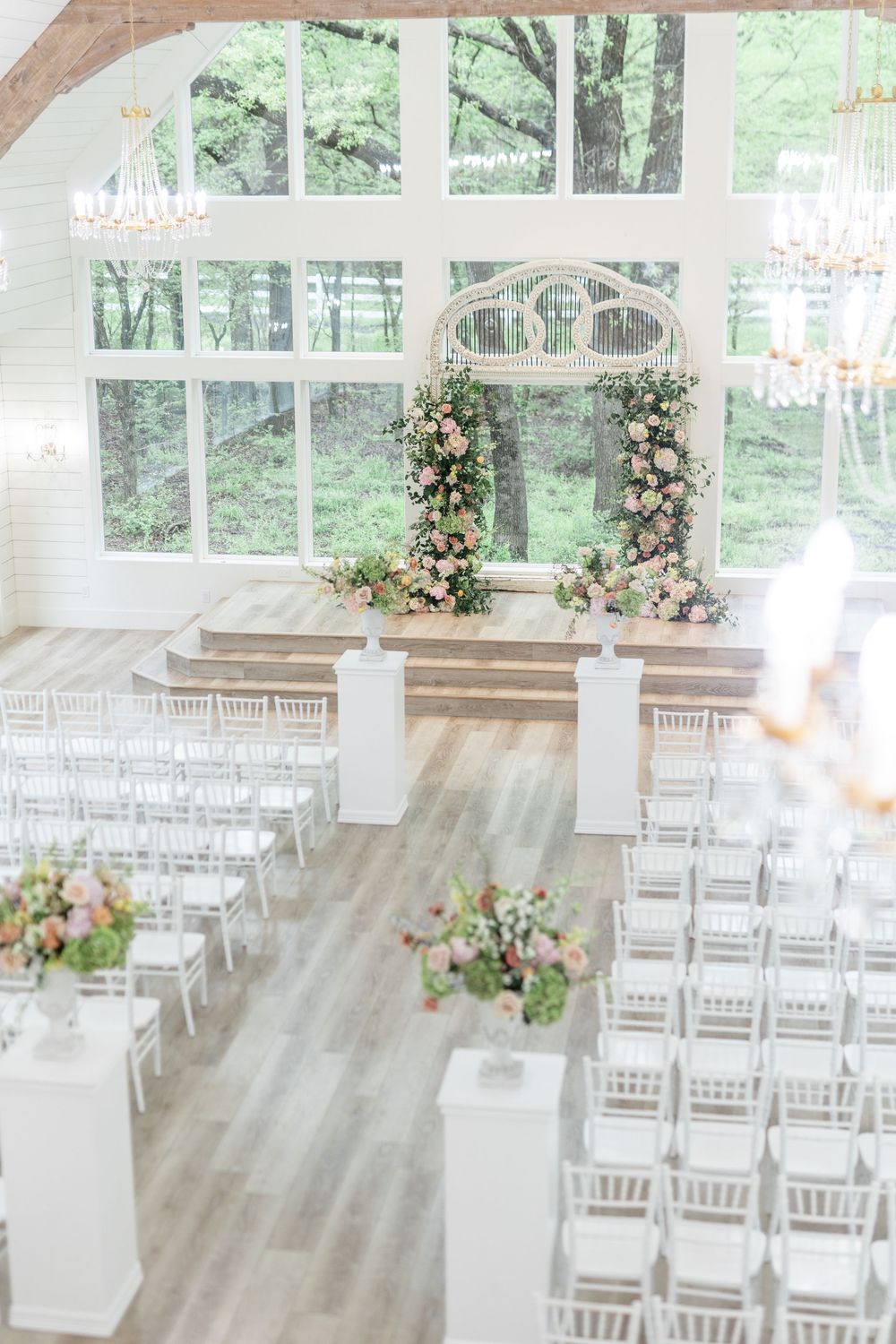 Elegant wedding chapel with white Chiavari chairs, floral arch, and large windows overlooking lush greenery.