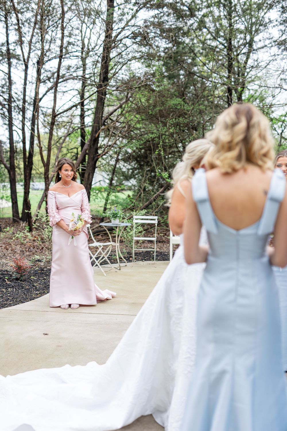 Bride and bridesmaid in light blue dress watch woman in pink gown approach on outdoor pathway.