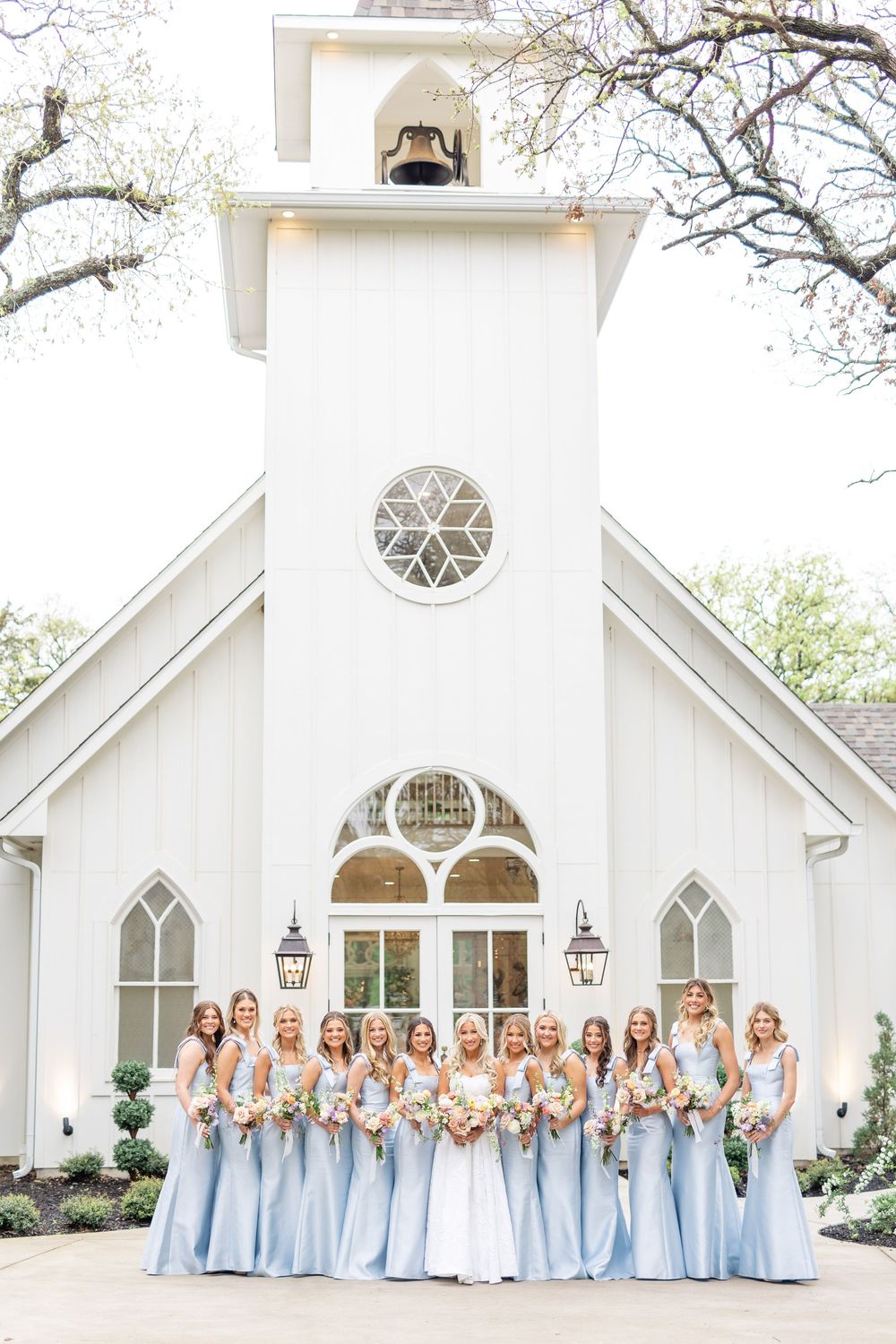 Bride in white gown poses with large bridal party in matching light blue dresses outside white chapel.