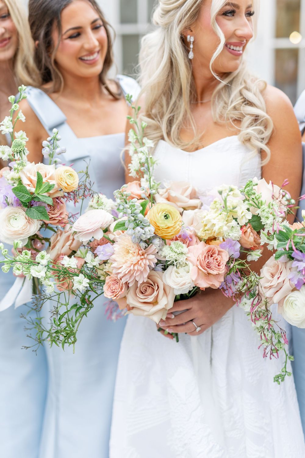Bride and bridesmaids in light blue dresses holding colorful pastel floral bouquets with roses and ranunculus.