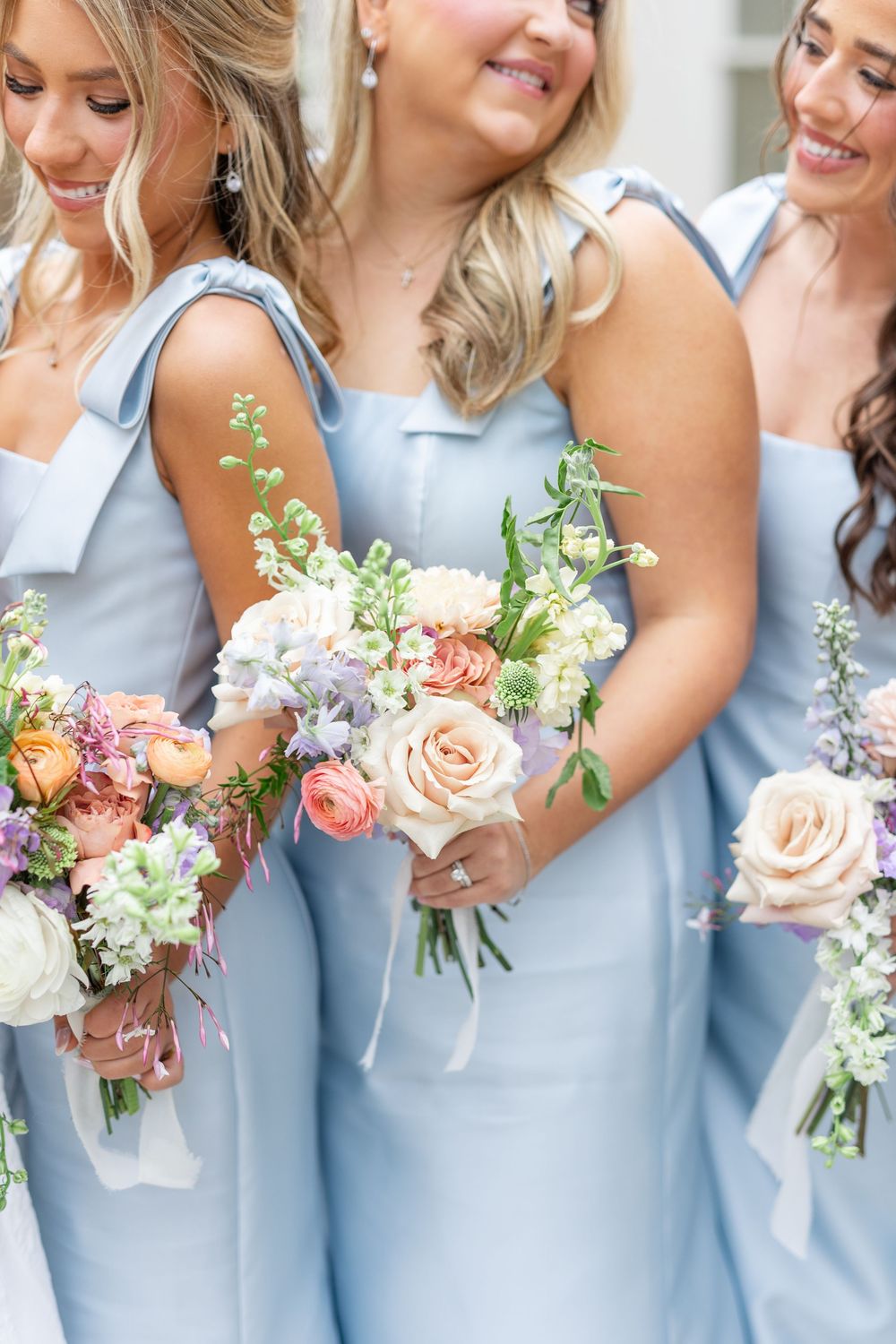 Bridesmaids in light blue dresses smiling and holding pastel bouquets with roses and wildflowers at wedding.
