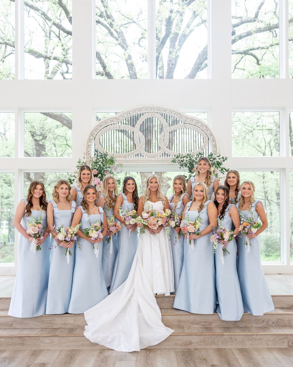Bride in white gown surrounded by bridesmaids in light blue dresses holding pastel bouquets indoors.