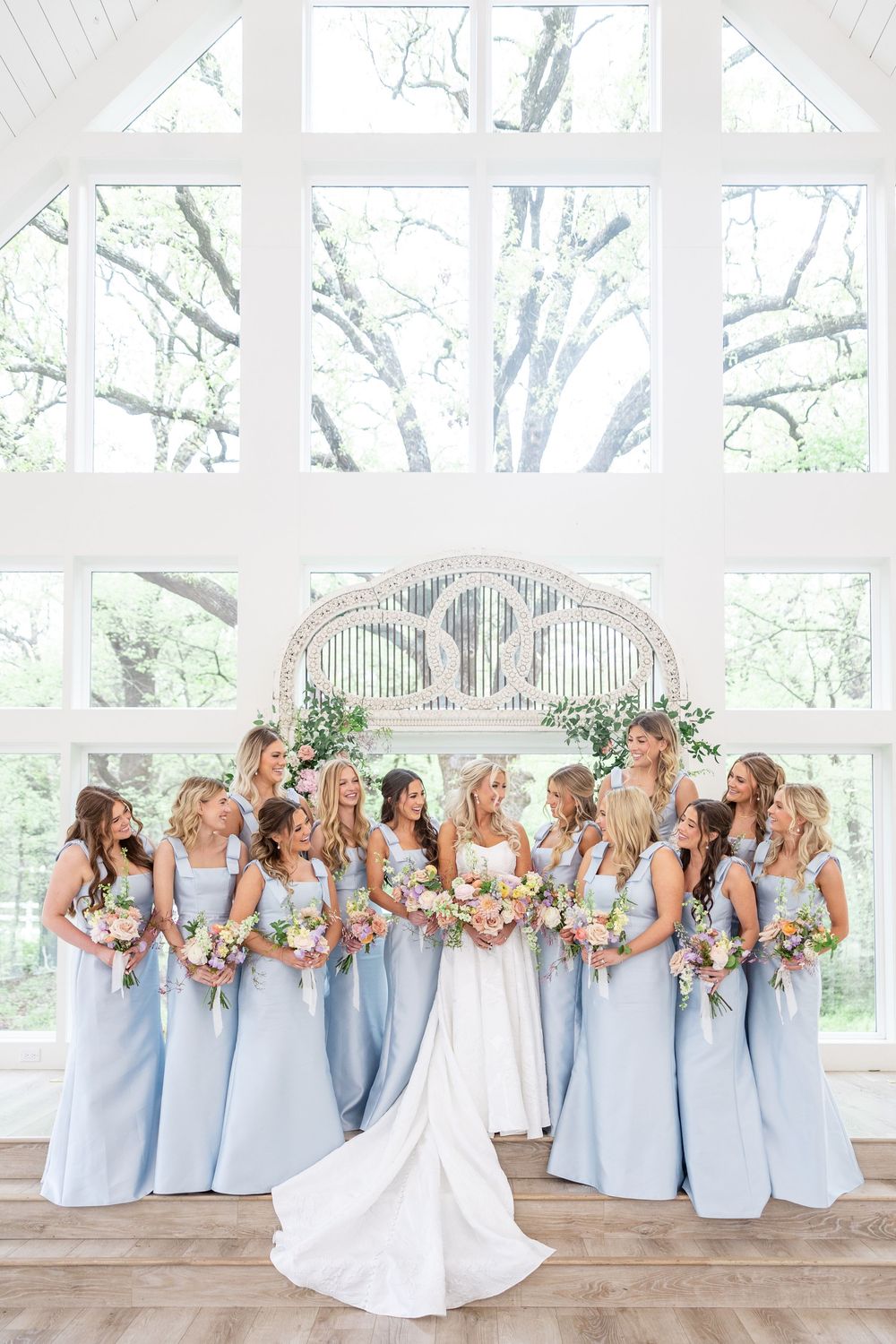 Bride surrounded by bridesmaids in light blue gowns holding colorful bouquets in a bright white chapel.