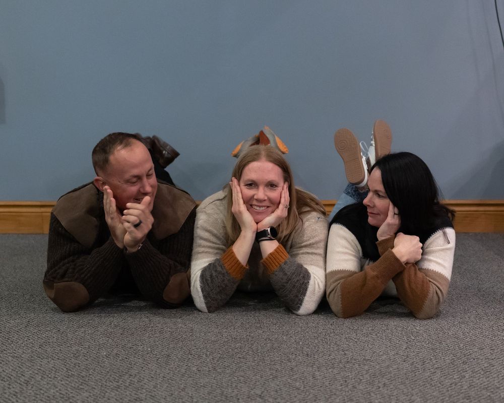 Three people lying on gray carpet with their chins resting on their hands, posed casually for a photo.