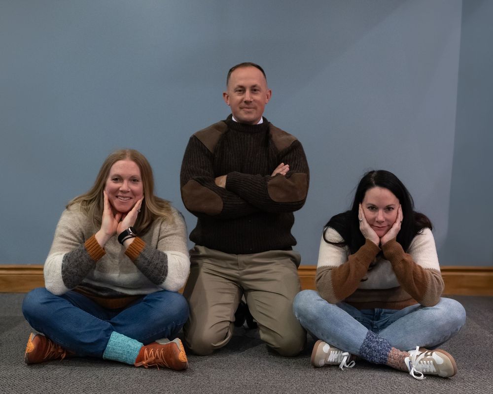 Three people pose casually on the floor against a blue wall, sitting and leaning in relaxed positions.
