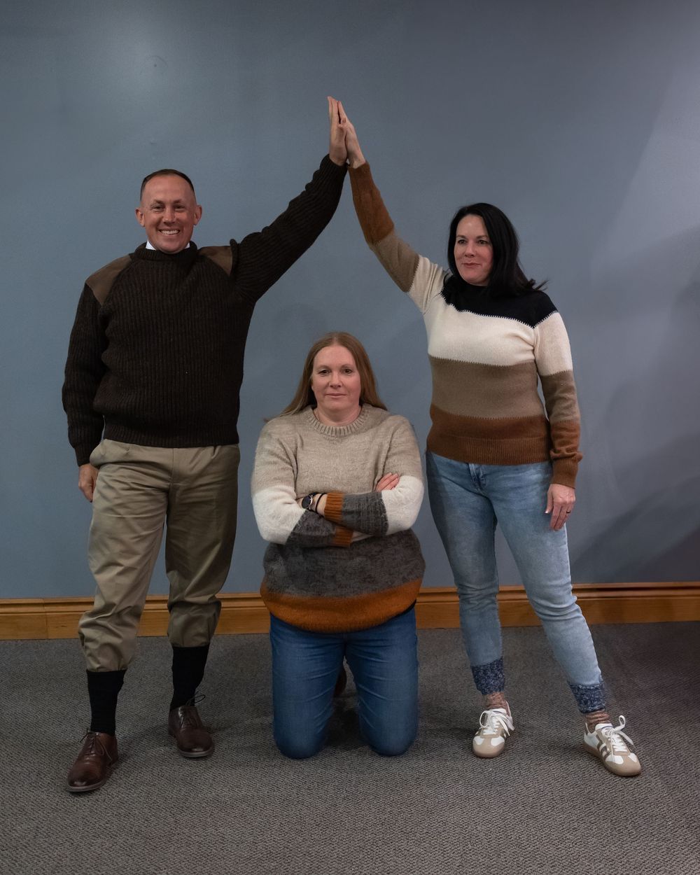 Three people in sweaters posing against a gray wall, two standing and one kneeling with raised arms in celebration.