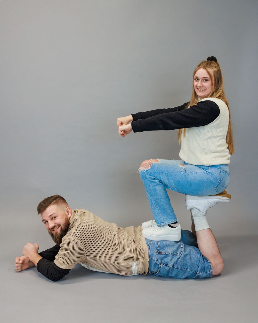 A playful couple poses together against a gray backdrop, with one person resting atop the other in a lighthearted pose.
