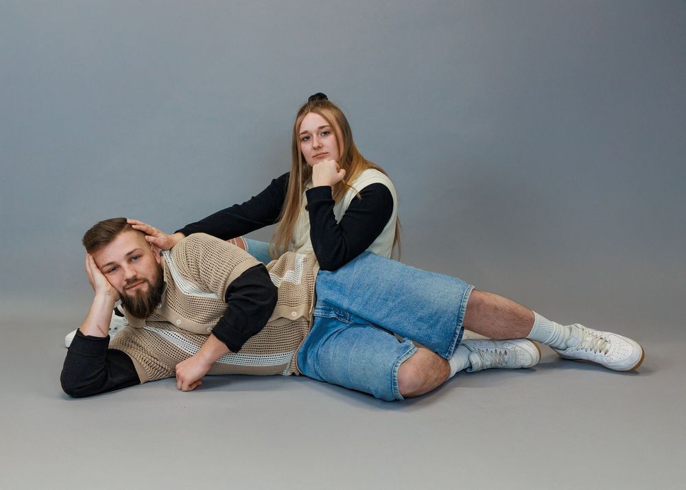Two people in casual attire posing playfully on a gray studio floor in matching black and beige outfits.