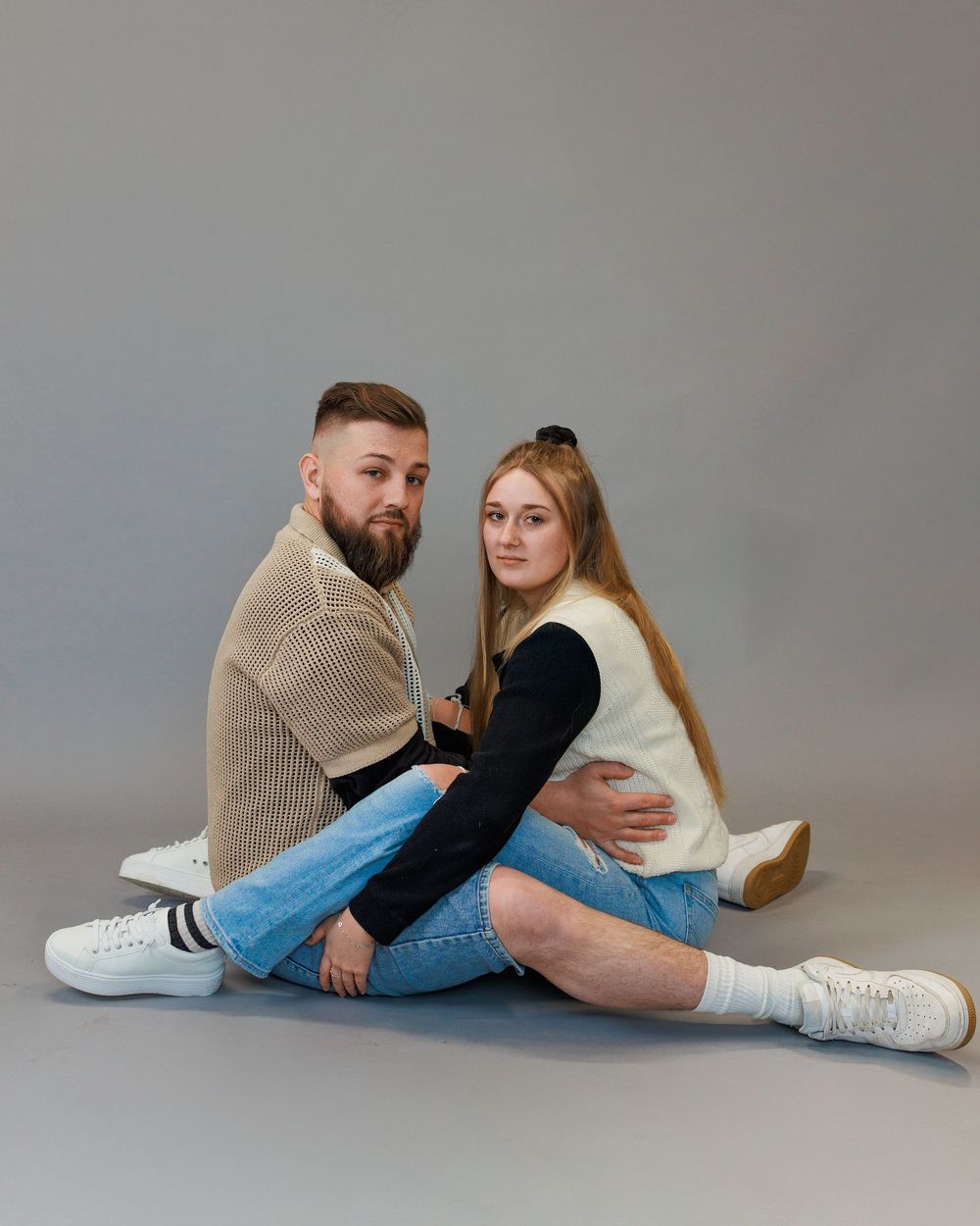 A couple in casual attire sits close together on the floor against a gray studio backdrop.