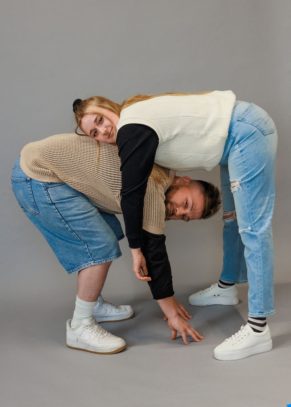 Two people pose playfully in casual clothing with one person bending over while the other leans on their back on a gray background.