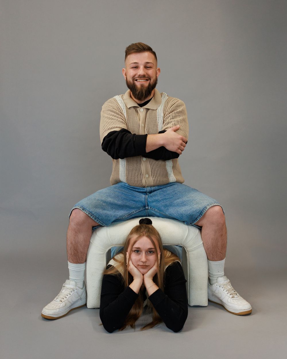 Two people posing for fun creative portrait on plain backdrop with knit sweater and denim shorts.