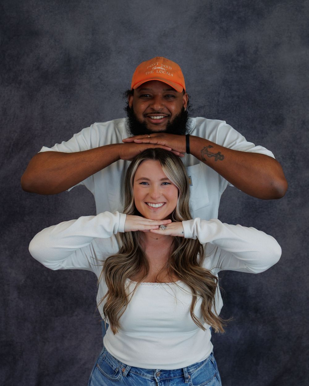 Two people in white shirts pose in a staggered formation making hand frames against a dark backdrop.