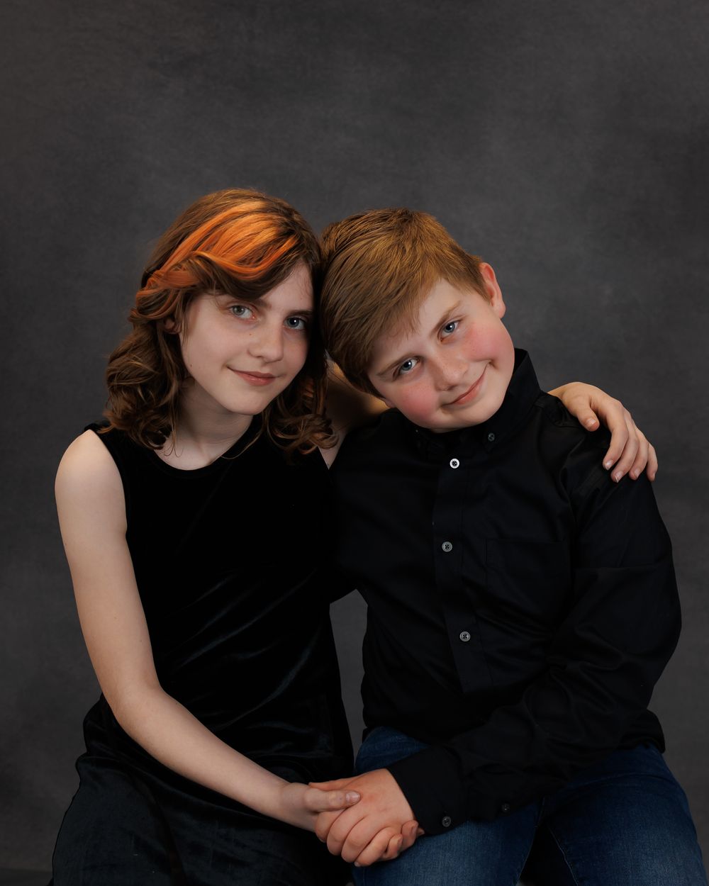 Studio portrait of a young boy and girl in formal black attire sitting close together against a dark backdrop.