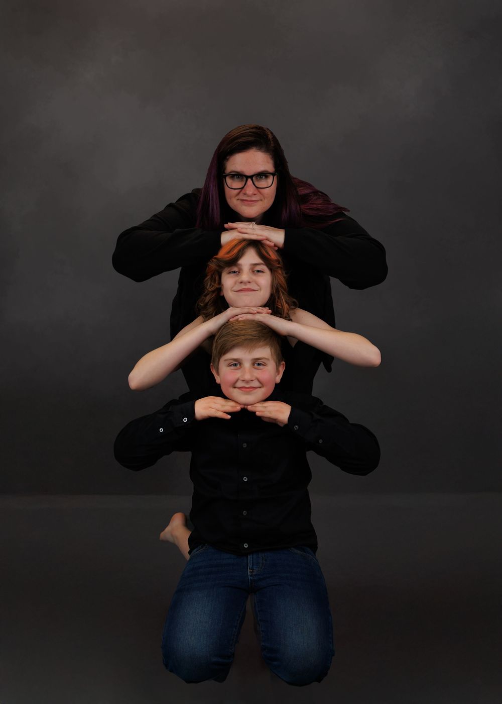 Three individuals in black shirts pose together in a creative stacked formation against a dark gray backdrop.