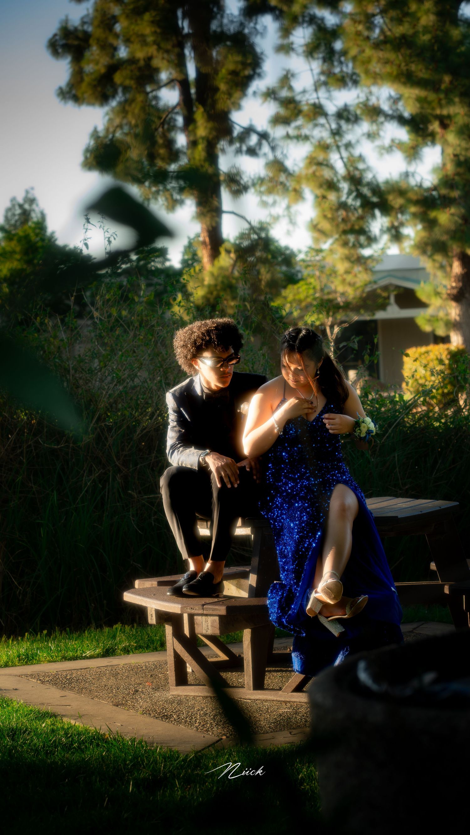 A couple sits together on a wooden bench in a garden setting, with the person in a sparkling blue dress glowing in sunlight.