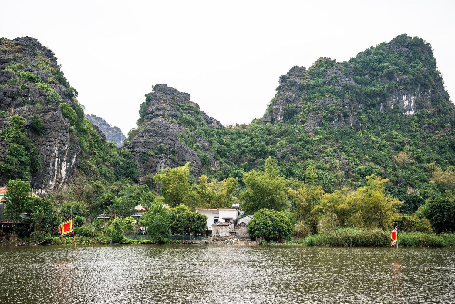 Dramatic limestone karst mountains rise above a tranquil lake with traditional buildings nestled at their base in Vietnam.