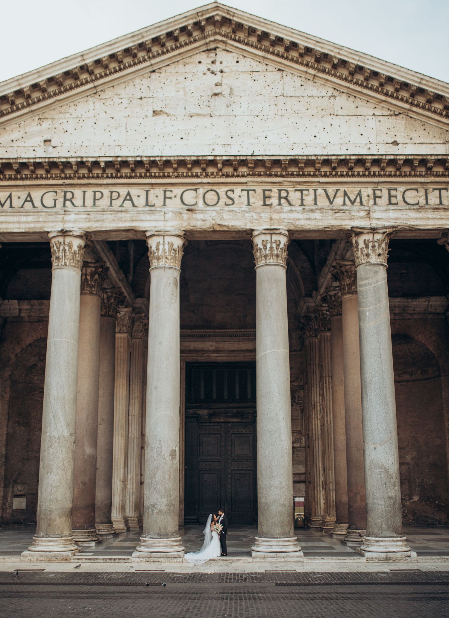 A couple in wedding attire stands beneath the grand columns of the historic Pantheon in Rome, Italy.
