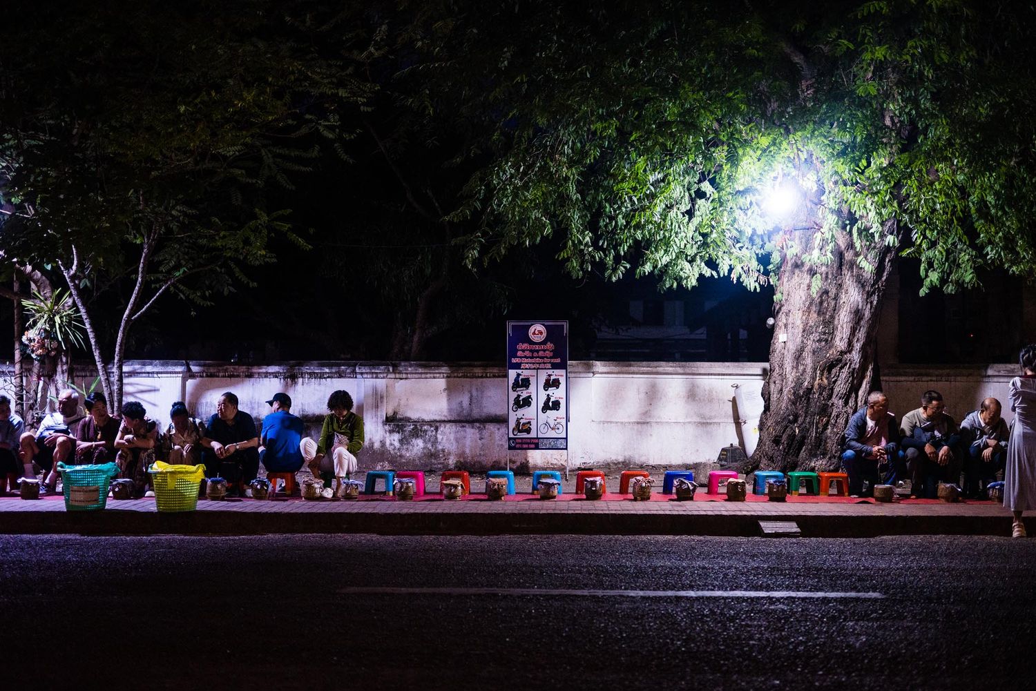 A dark nighttime scene shows a row of people sitting on benches illuminated by spotlights in a park setting.