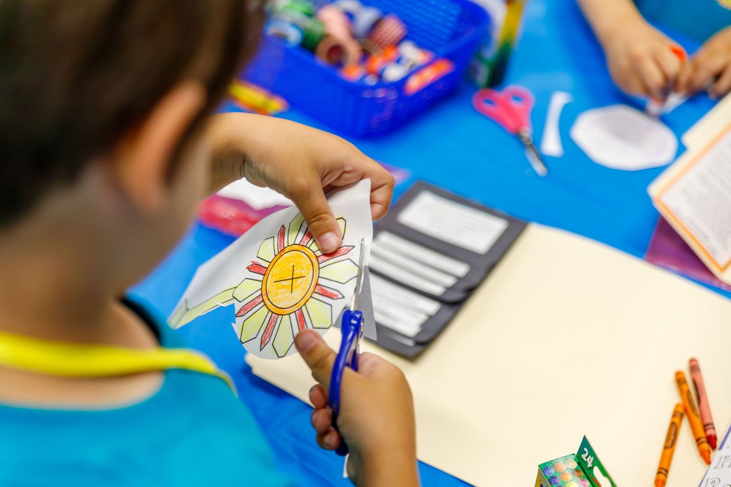 Student uses colored markers to draw a bright sun design during an art activity at a classroom desk.