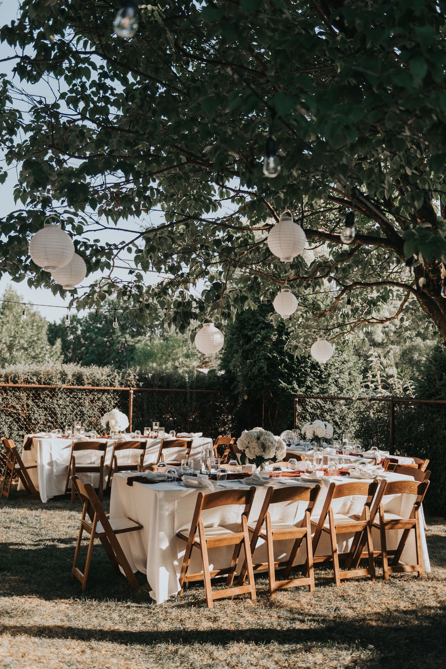 Rustic outdoor wedding reception setup with wooden chairs, white tablecloths, and hanging balloon decorations under trees.