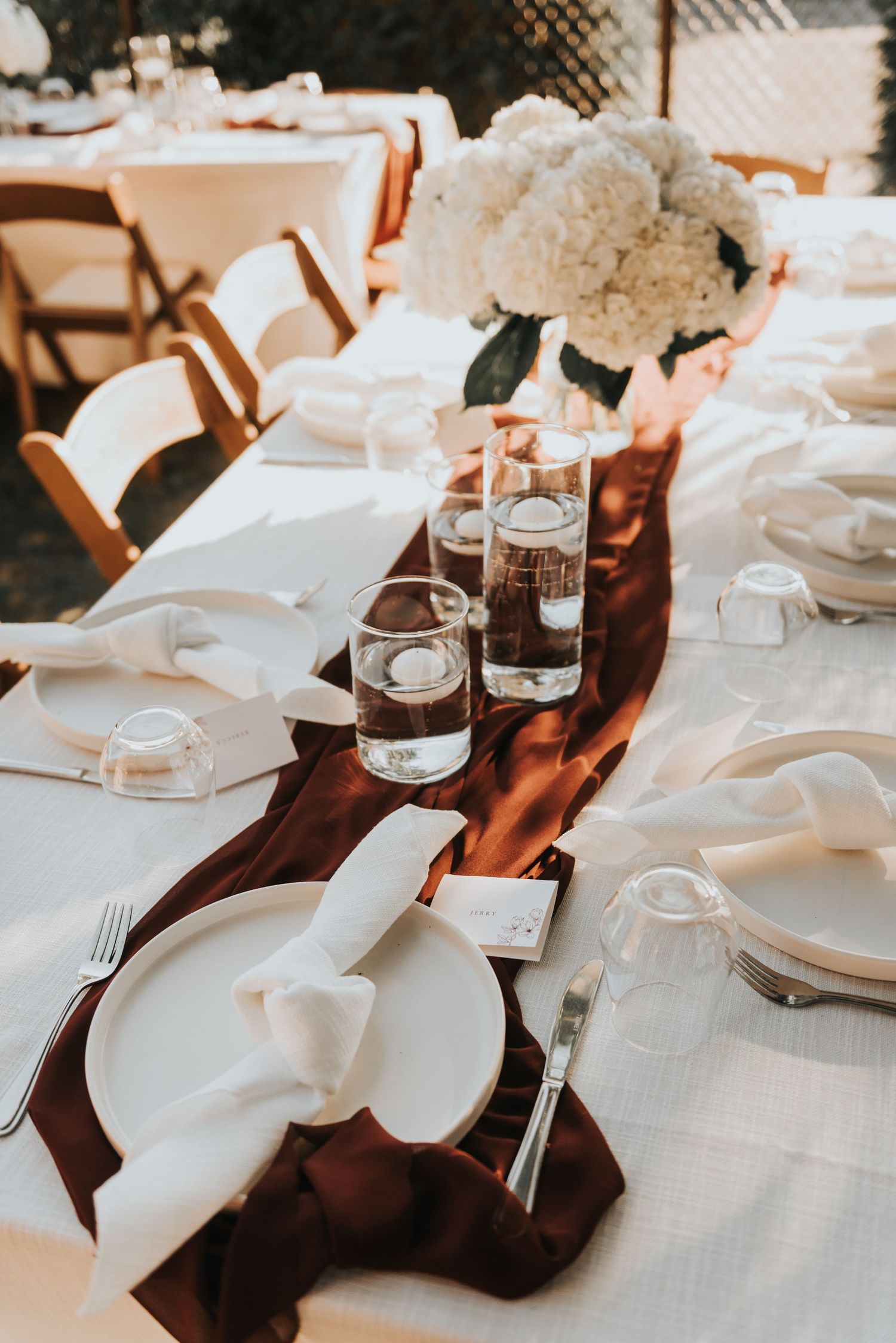 Elegant wedding reception table settings with white linens, burgundy napkins, and glass votive candle holders.