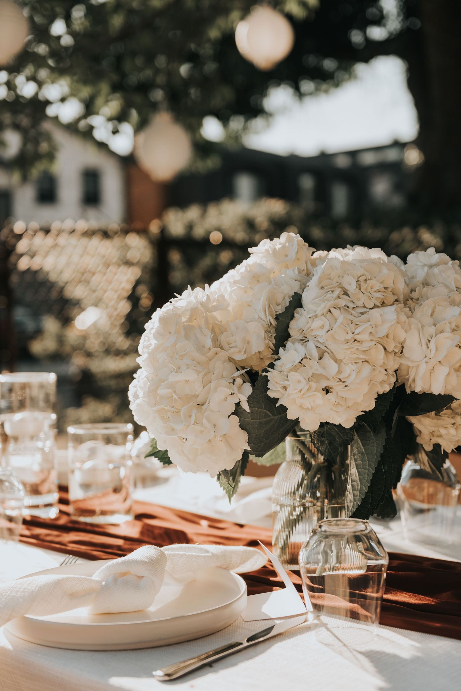 White hydrangea centerpieces in glass vases adorn an elegant outdoor table setting with white plates and warm lighting.
