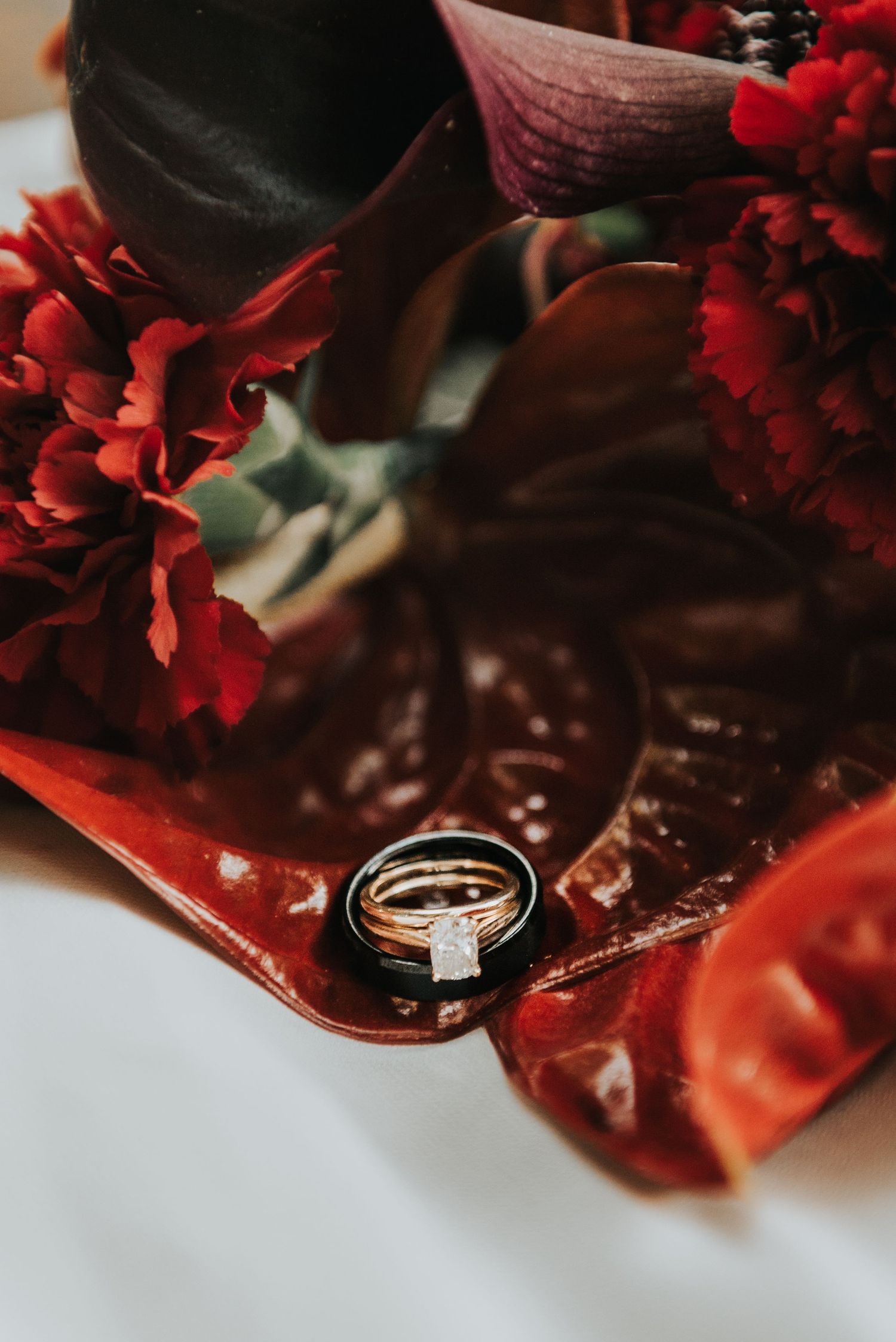 Diamond engagement ring nestled among red flowers and dark petals on a reflective surface with moody lighting.